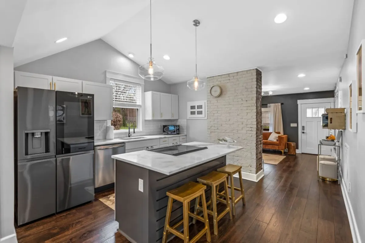 This image features a welcoming kitchen with an island and seating, set against a backdrop of a brick accent wall and a window letting in natural light. The space is designed for gatherings, casual dining, and entertaining friends, with a vibe that encourages group interaction and easy mornings after a night out.