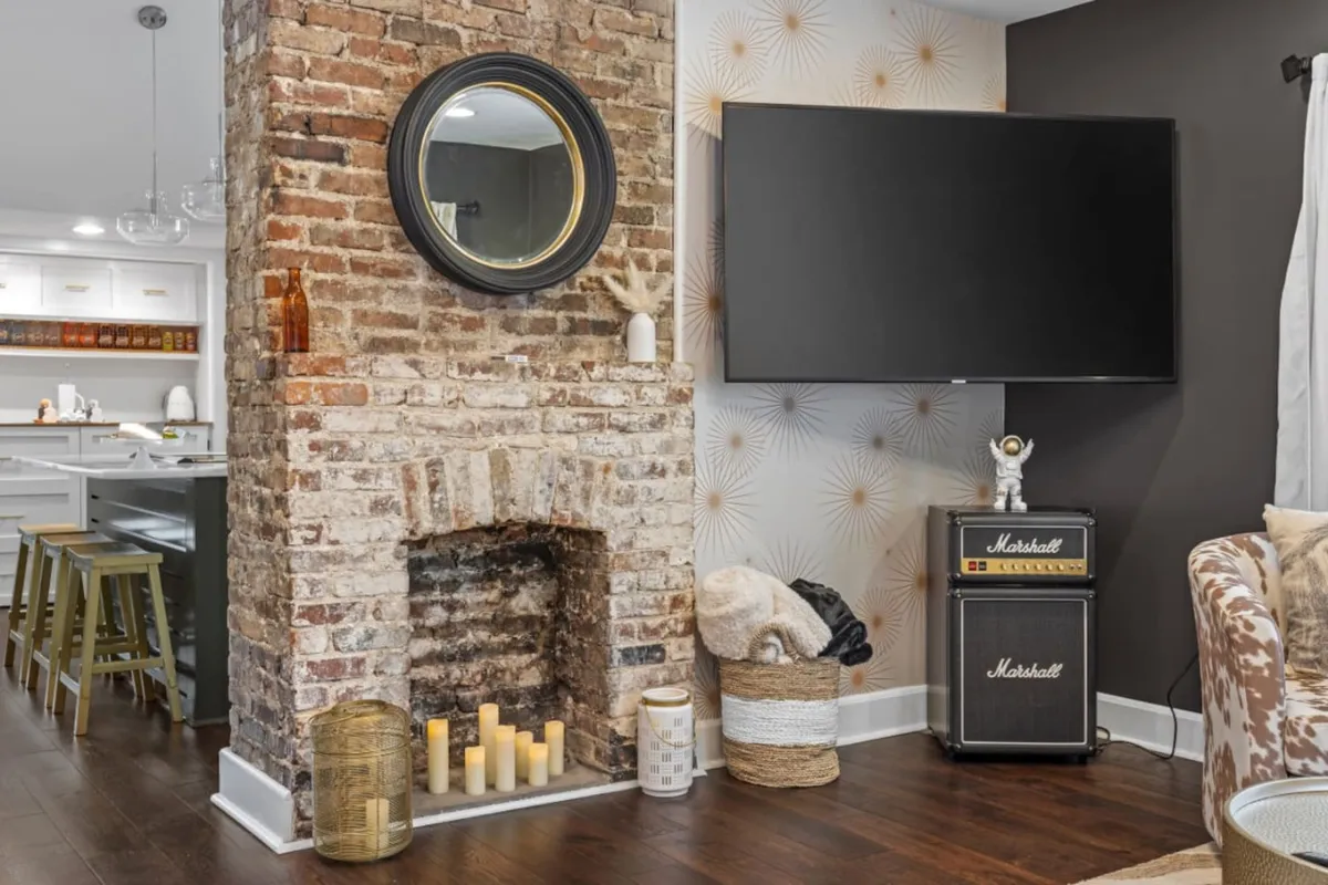 The photo features a cozy living room centered around a modern brick fireplace, a round mirror, television, and a Marshall amplifier. The space invites relaxation, movie nights, and casual family gatherings.