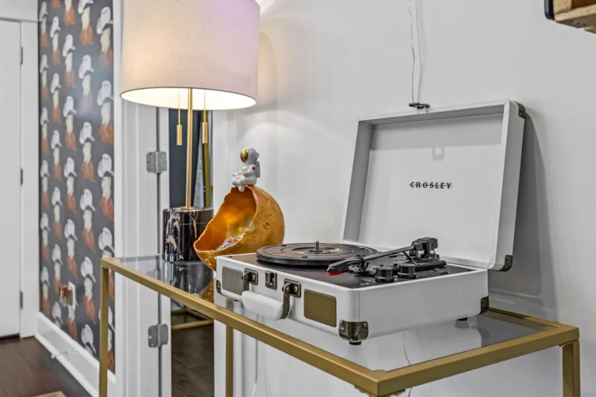 A cozy interior scene featuring a stylish record player, a decorative lamp casting a warm glow, and vibrant wall art, all arranged atop a thoughtfully curated sideboard. The space exudes an inviting, nostalgic mood, perfect for relaxing or sharing music together.