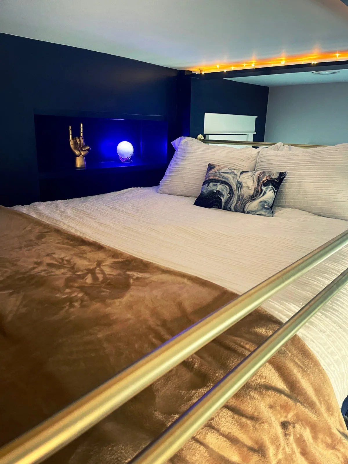 A serene bedroom scene featuring a bed with plush bedding, a glowing orb light on the bedside, a golden hand sculpture, and a decorative pillow. The overall mood is calming and restful, with thoughtfully chosen décor elements that add an artistic, boutique feel.