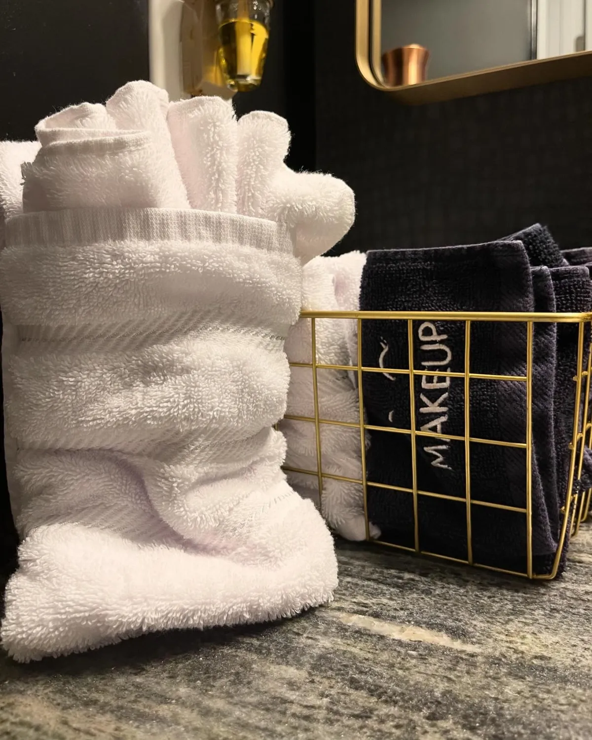 A serene bathroom scene featuring rolled white towels, dark towels neatly arranged in a gold wire basket, and a mirror reflecting the inviting ambiance. The soothing and organized setup suggests a focus on comfort and thoughtful amenities.