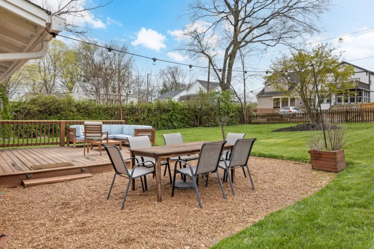 This exterior scene highlights a wooden deck with an outdoor dining table, a comfortable seating area, and a lush green lawn. The area appears perfect for afternoon relaxation, family gatherings, or evening celebrations. The scene is bathed in warm, natural light, evoking feelings of casual enjoyment and togetherness.